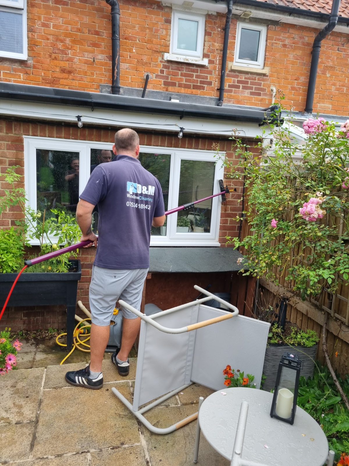 Matthew in a D&M polo shirt cleaning a back window with a water-fed pole, in a garden with roses