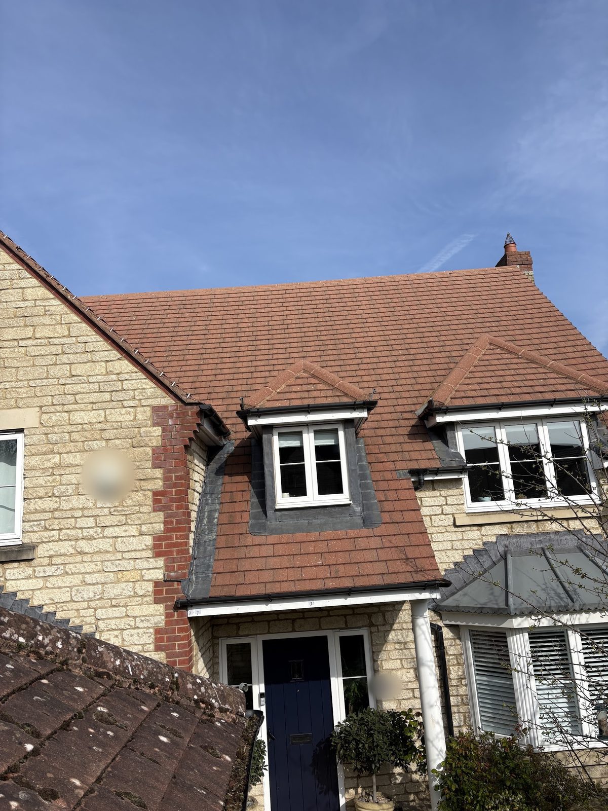 A Cotswold stone house front with a dormer window and a navy front door, seen from a mossy roof